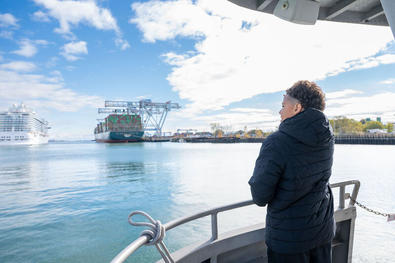 The image shows a person on a boat looking out at a harbor. In the background, there is a large ship and a container ship. The sky is blue with some clouds. The person is wearing a dark jacket. The overall scene is bright and sunny.