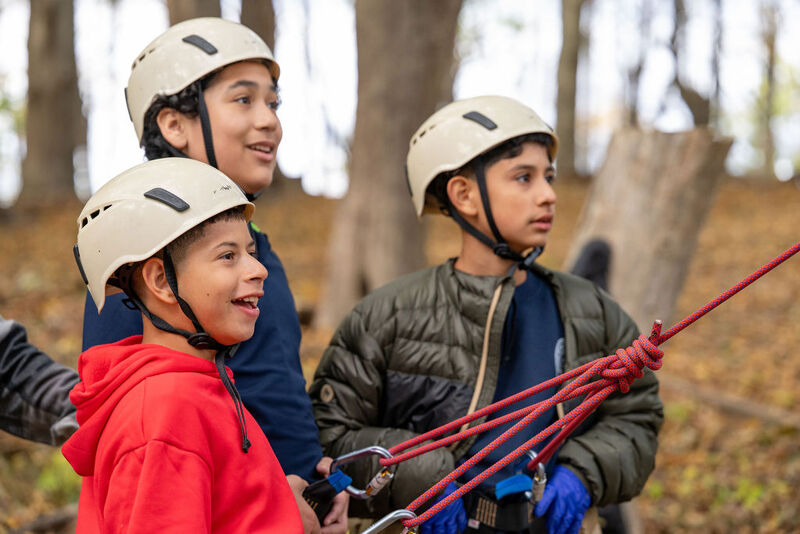 The image shows three young boys wearing helmets, likely preparing for an outdoor activity like a ropes course or climbing. They appear to be looking at something with anticipation. The boy in the red hoodie is smiling. The background suggests a wooded area.