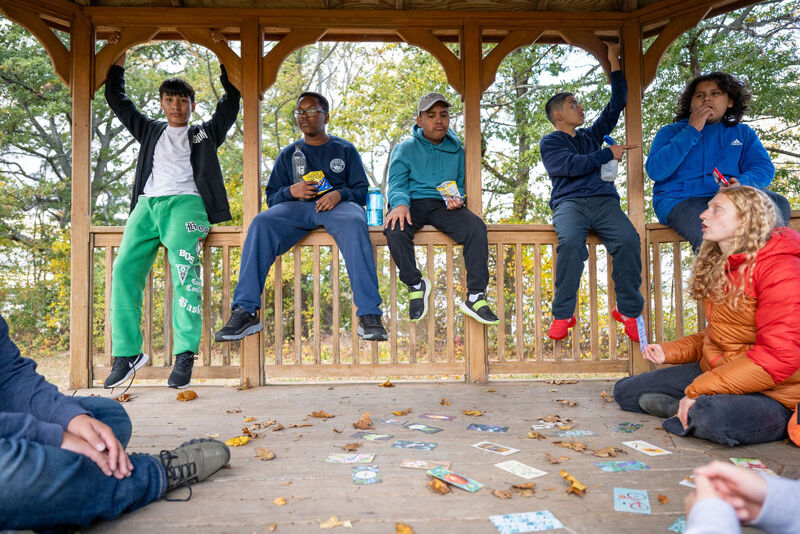 The image shows a group of young people in a gazebo. Some are sitting on the railing, while others are standing or sitting on the floor. They appear to be playing a game, with cards scattered on the floor. The atmosphere seems relaxed and social.