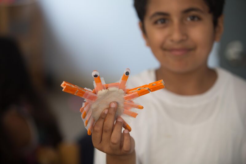 The image shows a young boy holding a craft project resembling a crab. The crab is made from clothespins, googly eyes, and other craft materials. The boy is smiling and looking at the camera, suggesting pride in his creation. The background is blurred, keeping the focus on the boy and his craft.