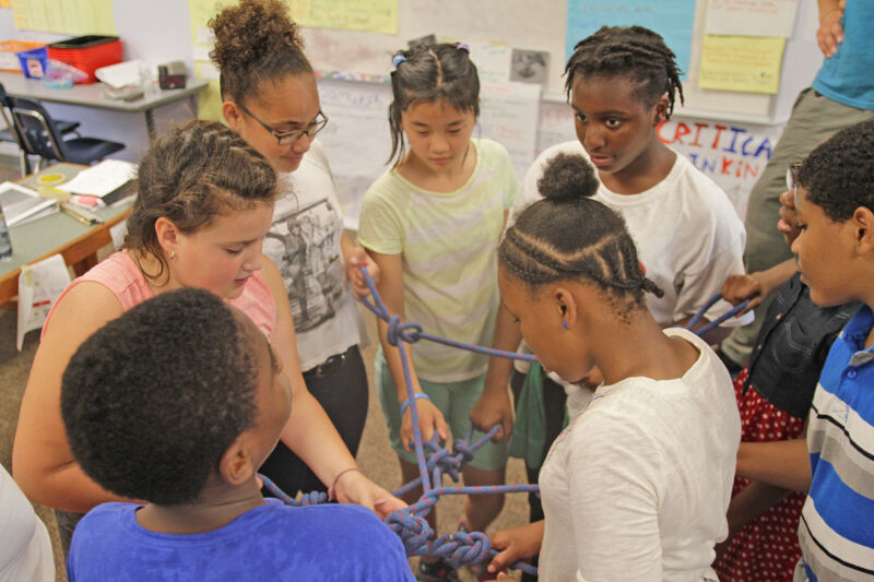The image shows a diverse group of children working together on a collaborative project. They are gathered around a rope structure, likely engaged in a problem-solving activity. Their focused expressions suggest active participation and teamwork. The setting appears to be a classroom, with posters and other educational materials visible in the background.