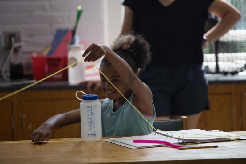In the image, a young girl is playing with a string, her face lit up with a smile. She is sitting at a table with a water bottle and some papers. An adult is standing in the background. The scene appears to be indoors, possibly in a kitchen or a similar space.