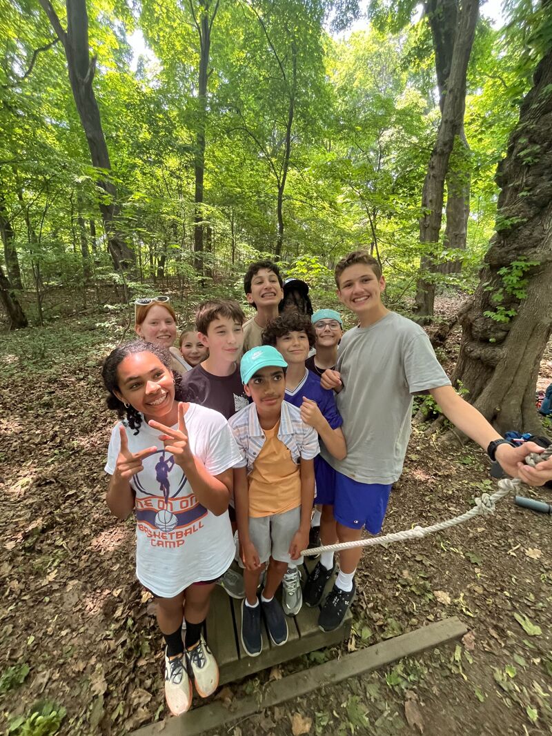 The image shows a group of young people posing for a photo in a forest. They are standing on a wooden structure, and some are smiling at the camera. The trees in the background create a natural setting. It seems like they are enjoying an outdoor activity.