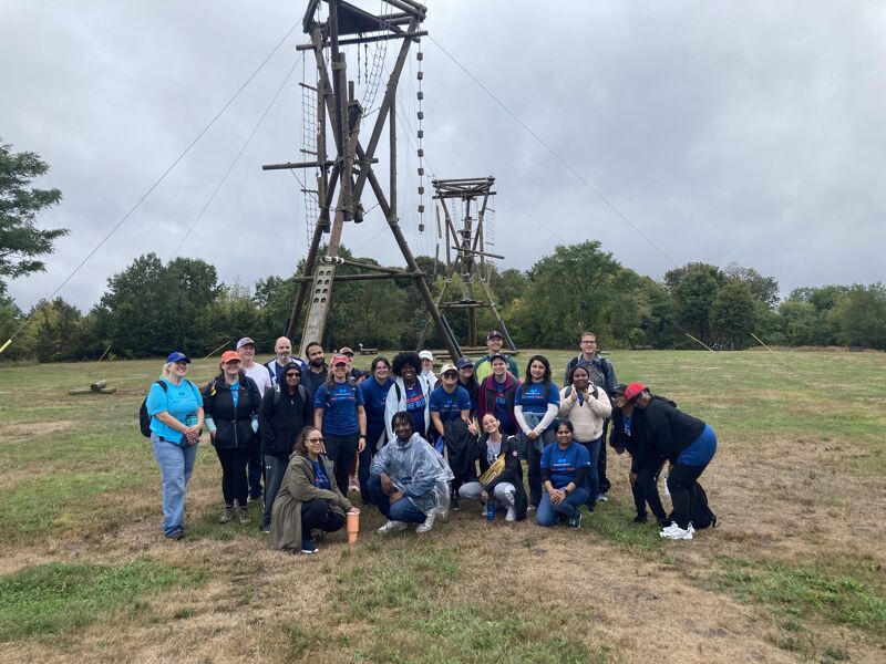 The image shows a group of people posing for a photo in front of a wooden structure, likely an adventure course. The people are of diverse ages and ethnicities, dressed in casual clothing. The setting appears to be a grassy field on a cloudy day. The structure has ropes and platforms, suggesting an outdoor activity or team-building exercise.