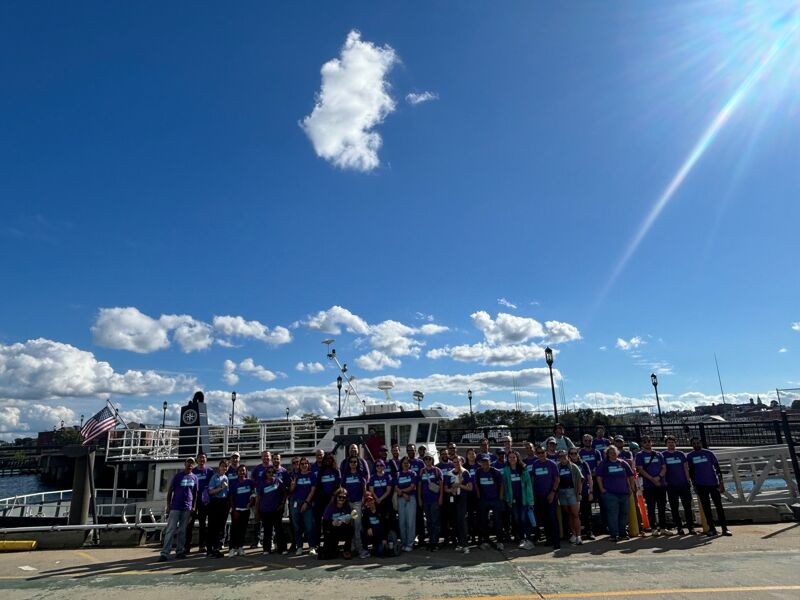 The image shows a large group of people posing for a photo outdoors on a sunny day. They are standing in front of a boat. The sky is blue with some clouds. Most of the people are wearing purple shirts. The overall scene suggests a group outing or event.