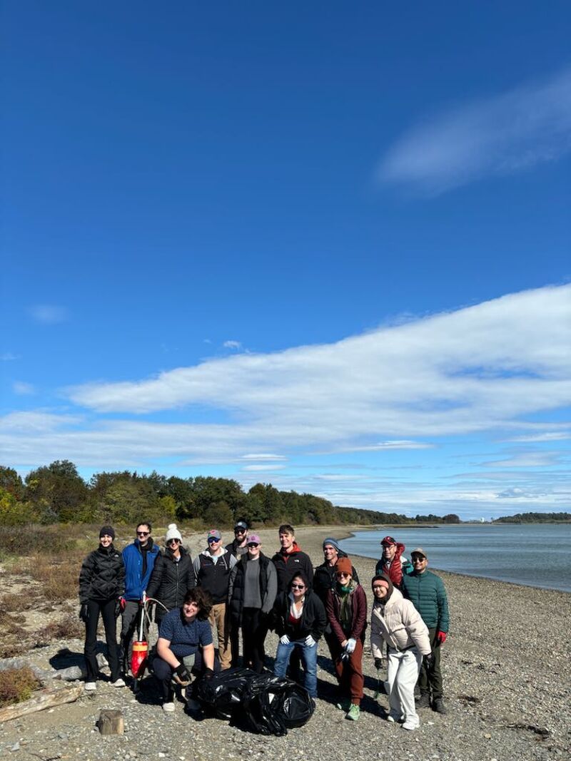 The image shows a group of people on a beach. They appear to be volunteers, possibly participating in a cleanup event. Some are holding trash bags, and the overall scene suggests an effort to preserve the environment. The sky is blue with some clouds, and the weather seems pleasant.