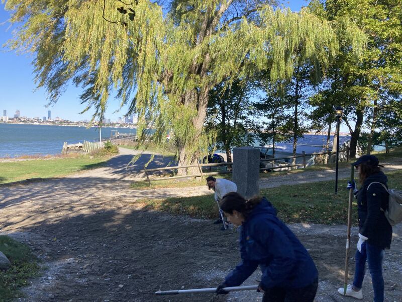 The image shows a group of people cleaning up an outdoor area. They are using tools to pick up trash or debris. In the background, there is a large tree, a body of water, and a city skyline. The weather appears to be sunny.