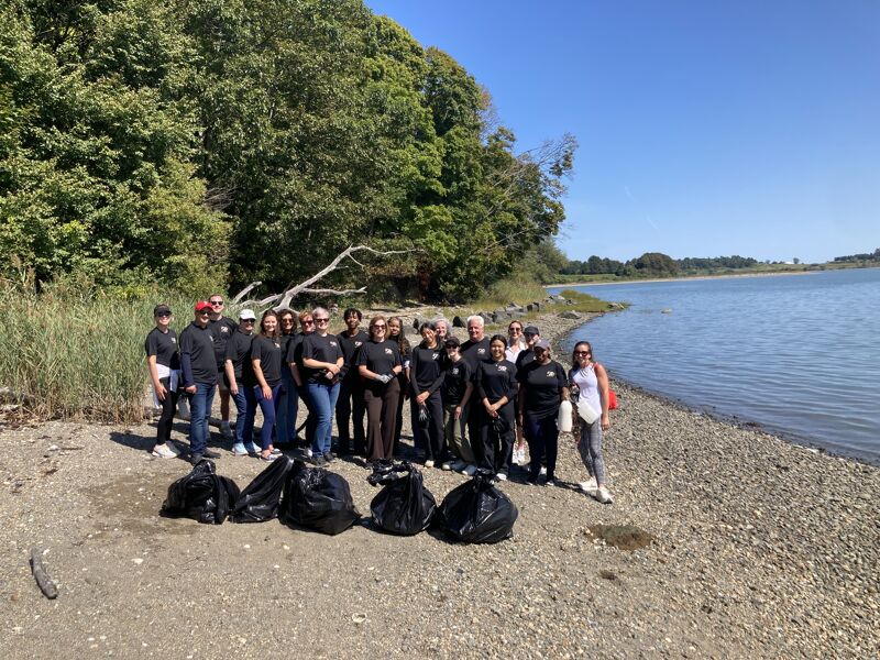 A group of people stands on a beach next to black trash bags. They appear to have been cleaning up the area. The sky is blue, and there is a body of water in the background. The people are wearing dark-colored clothing.