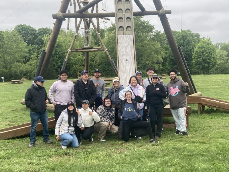 The image shows a group of people posing for a photo in front of a wooden structure. The structure appears to be part of an outdoor activity or adventure course. The people are of diverse ages and ethnicities, and they are all smiling and looking at the camera. The setting is a grassy field with trees in the background.