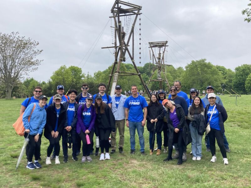 The image shows a group of people posing for a photo outdoors. They are standing in front of a wooden structure, likely part of an adventure course. Most of them are wearing blue shirts, and the overall atmosphere seems casual and friendly. The background includes trees and a cloudy sky.