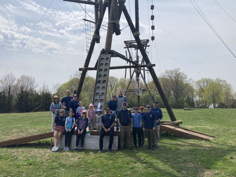 The image shows a group of people posing in front of a wooden structure, likely an obstacle course or climbing tower. The people appear to be a mix of adults and children, suggesting a recreational or team-building activity. The setting is outdoors on a grassy field, with trees and a cloudy sky in the background.
