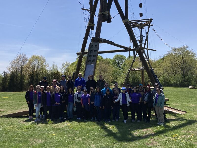 The image shows a group of people posing for a photo in front of a wooden structure, likely part of an adventure course. The people are of various ages and are dressed in casual attire. The setting appears to be a grassy field on a sunny day.