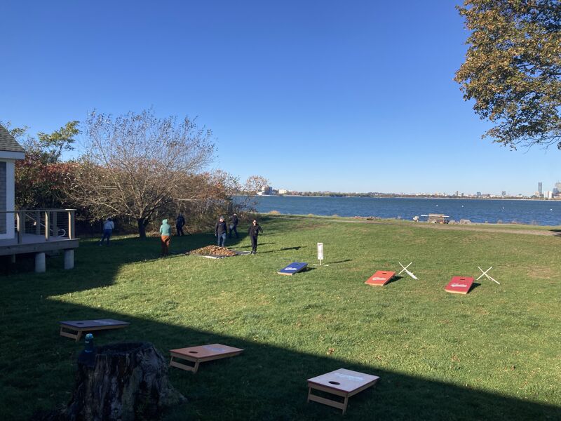 The image shows a sunny outdoor scene with a grassy area. There are several cornhole boards set up, suggesting a game is in progress or about to begin. People are scattered around the yard, and a body of water is visible in the background. The sky is clear and blue.