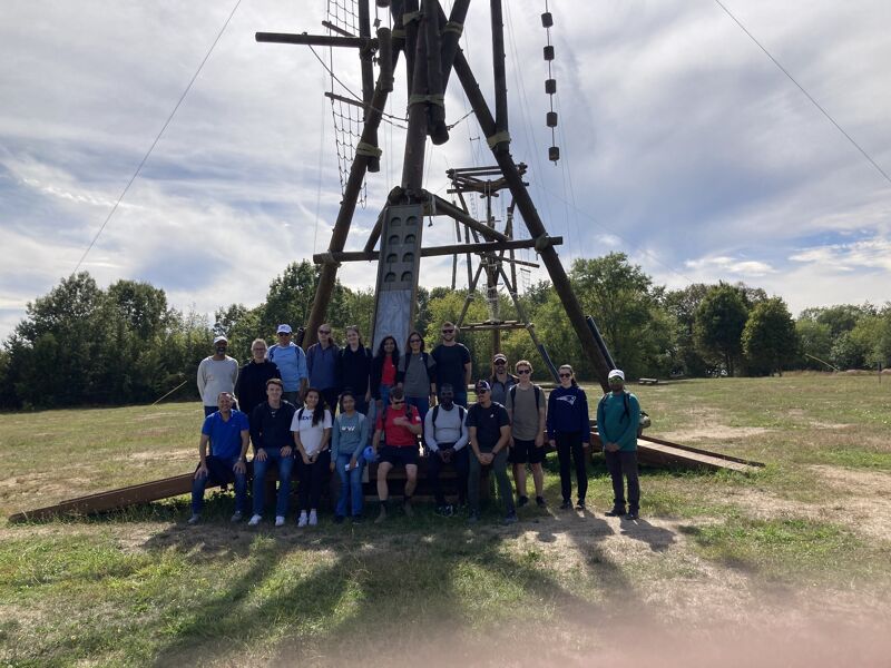 The image shows a group of people posing in front of a wooden structure, likely part of an adventure course. The structure has ropes and platforms, suggesting it's used for climbing or other activities. The people are diverse in age and attire, indicating a group outing or team-building event. The setting is outdoors, with trees and a cloudy sky in the background.