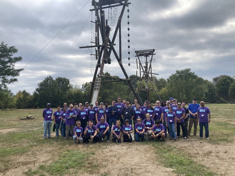 The image shows a large group of people posing for a photo outdoors. They are wearing matching purple shirts and are in front of a wooden structure with ropes and platforms, likely an obstacle course. The sky is overcast, and the setting appears to be a field or park.