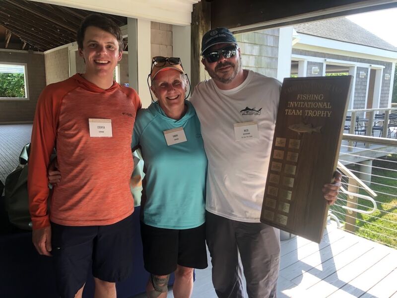 The image shows three people posing for a photo. They are holding a trophy that says "Fishing Invitational Team Trophy". They are all smiling and appear to be happy. The setting seems to be outdoors, possibly at a fishing event.