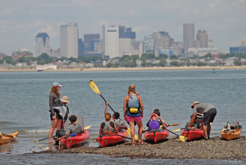 The image shows a group of people preparing to kayak on a beach. They are near the water's edge, with kayaks in the foreground. In the background, there is a city skyline. The sky is overcast. The people appear to be getting ready for a kayaking trip.