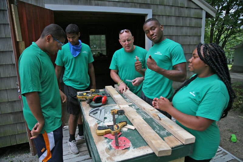 The image shows five people, all wearing green shirts, gathered around a workbench. They appear to be working on a project, possibly carpentry, with tools and wood visible. The setting seems to be outdoors, near a wooden structure. The expressions on their faces suggest they are focused on the task at hand.
