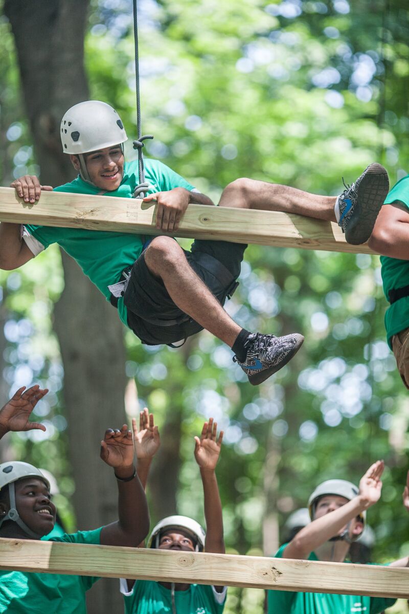 The image shows a young man on an obstacle course, suspended between wooden beams. He is wearing a helmet and a green shirt, with others below reaching up to him. The setting appears to be outdoors, with trees in the background, suggesting a camp or recreational activity.