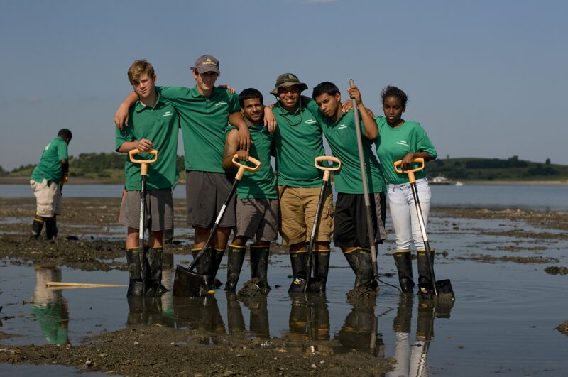 The image shows a group of people standing in the water, posing for a photo. They are wearing matching green shirts and holding shovels. They appear to be working on a project together, possibly related to environmental conservation or cleanup. The background includes a body of water and a clear sky.