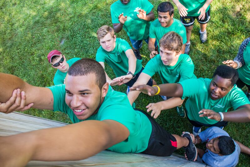 The image shows a group of young men in green shirts participating in an outdoor activity. One man is in the foreground, seemingly climbing or being supported by others. The others are reaching out to help. The scene suggests teamwork and cooperation. The setting appears to be a sunny day.