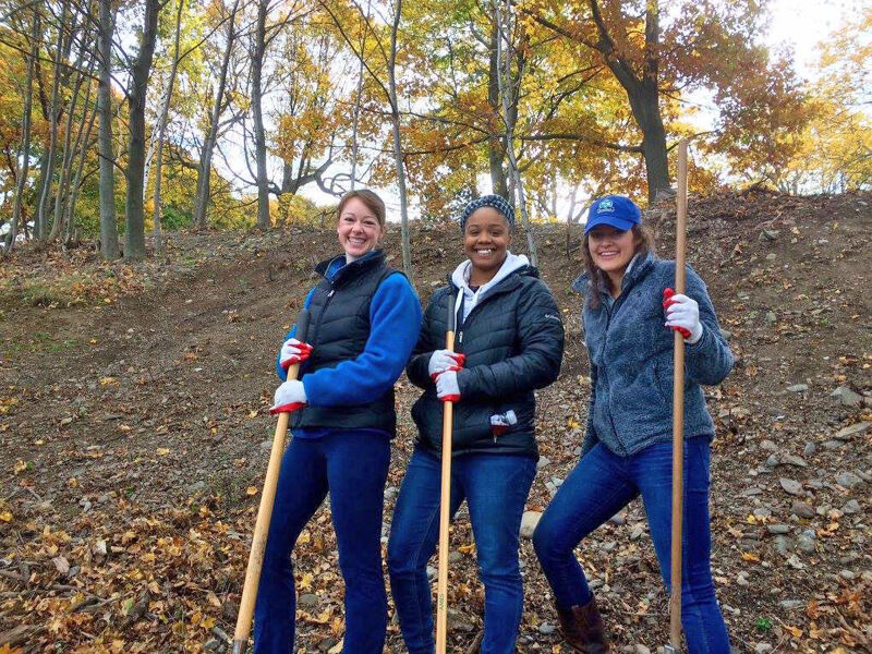 The image shows three women standing outdoors, each holding a rake. They are smiling and appear to be participating in a volunteer activity, possibly raking leaves. The background features trees with autumn foliage. The women are dressed in casual attire suitable for outdoor work.