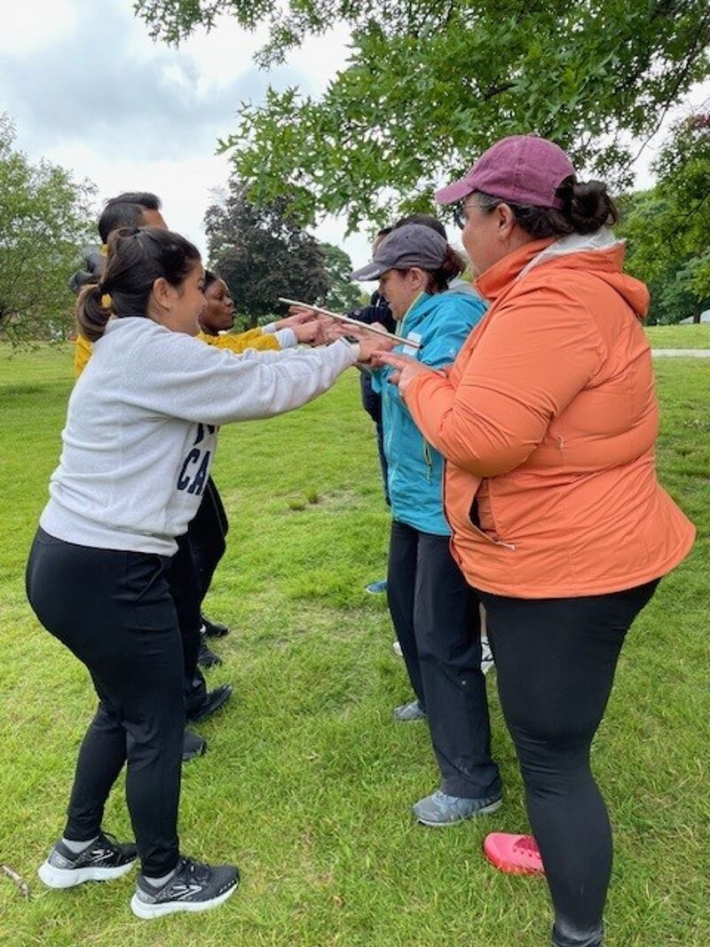 A group of people are participating in an outdoor team-building activity. They are standing in a grassy area with trees in the background, and they are all holding a long stick horizontally, with their index fingers supporting it. The activity appears to require coordination and communication as they work together to balance the stick.