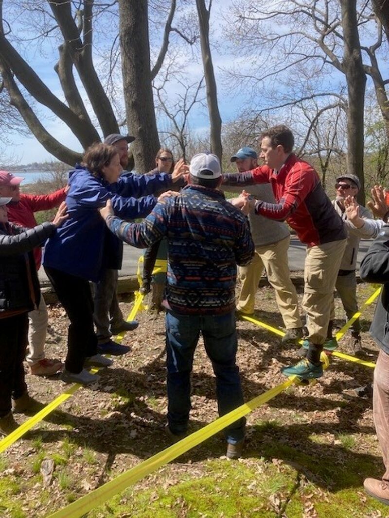 A group of people are playing a game outdoors, possibly a team-building exercise. They are standing in a circle, holding onto each other, with yellow tape forming a boundary or obstacle on the ground. The atmosphere appears energetic and collaborative, with participants actively engaged in the activity amidst a natural, wooded setting.