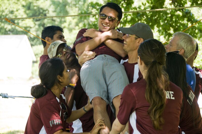 In the image, a group of people are holding up a man. The man is smiling and wearing sunglasses. They are all wearing matching maroon shirts. It looks like they are participating in a team-building activity outdoors. The background is blurred, suggesting a sunny day.