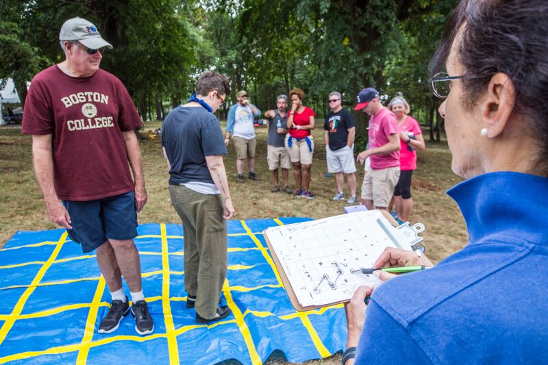 The image shows a group of people outdoors, likely participating in a team-building activity. A woman in the foreground is holding a clipboard and appears to be observing the activity. Several people are standing on a blue mat with yellow lines, while others are watching. The atmosphere seems relaxed and collaborative.