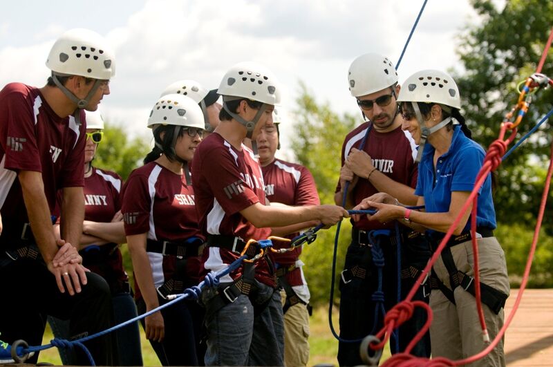 The image shows a group of people wearing helmets and harnesses, likely preparing for a ropes course or similar activity. They appear to be receiving instructions or checking equipment. The setting is outdoors, with trees and a cloudy sky in the background. The people are dressed in maroon shirts and blue shirts.