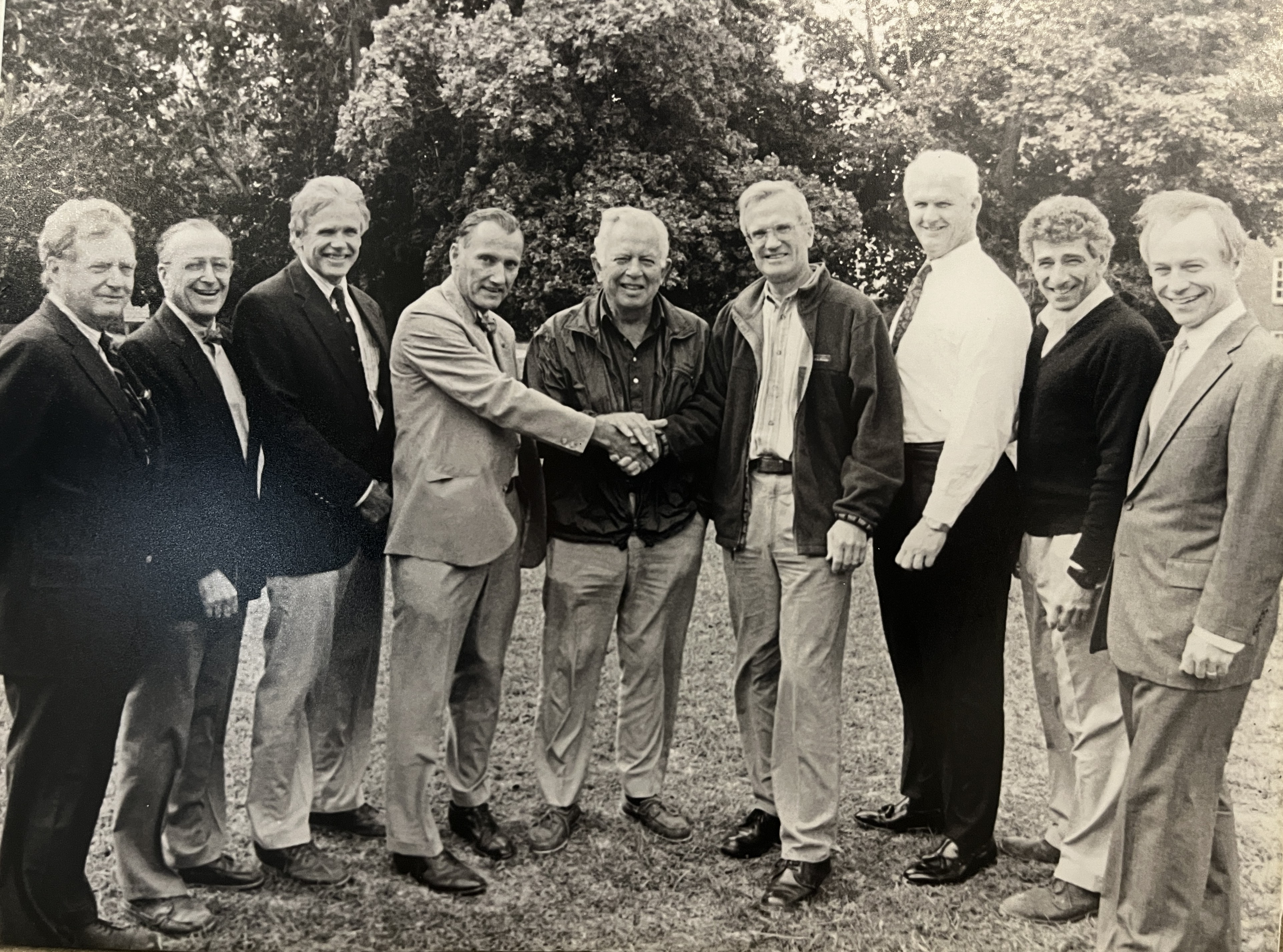 The image shows a group of men, likely in a business or formal setting, shaking hands. They are all dressed in suits or smart casual attire. The photo appears to be taken outdoors, possibly on a lawn, and the style suggests it might be an older photograph.