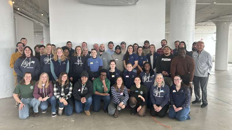 The image shows a large group of people posing for a photo indoors. They are of diverse ages and ethnicities, and most are wearing similar blue shirts with a logo. The background is a plain white wall and pillars. The overall impression is of a team or group gathering.