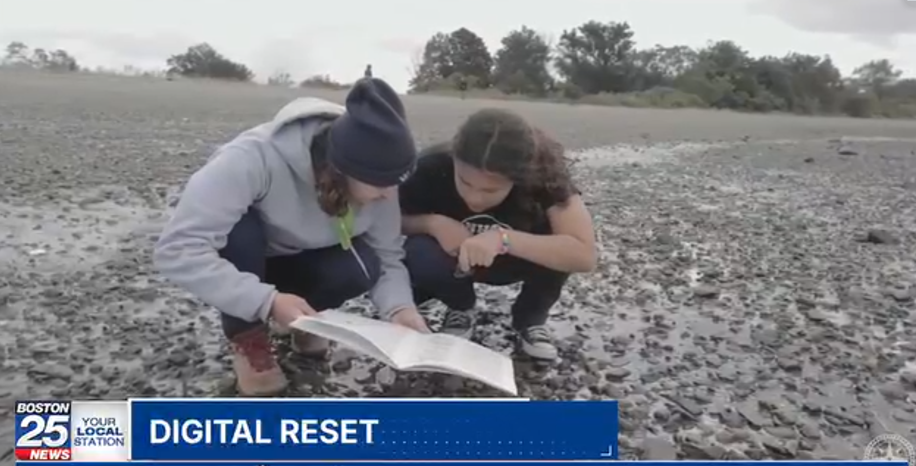 The image shows two people looking at a book on a rocky shore. They are both crouched down, examining the pages. The scene is set outdoors, with a body of water and some trees in the background. The bottom left corner of the image has a news station logo.