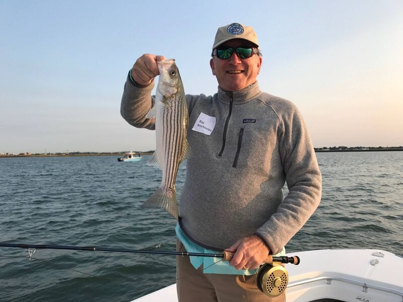 A man is holding a striped bass he just caught while on a boat. He is wearing a baseball cap, sunglasses, a gray fleece pullover, and khaki pants. The man is smiling and appears to be enjoying his fishing trip on a sunny day. The body of water looks calm with a small boat in the background.