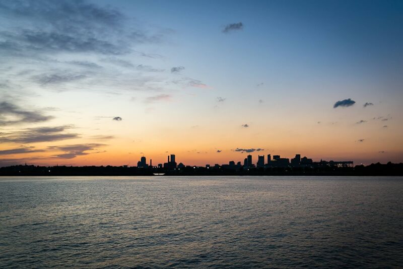 This image captures a serene sunset over a body of water, with the silhouette of a city skyline visible in the distance. The sky transitions from a deep blue at the top to warm hues of orange and yellow near the horizon, where the sun is setting. Wispy clouds add texture to the sky, and the calm water reflects the fading light, creating a peaceful and picturesque scene.