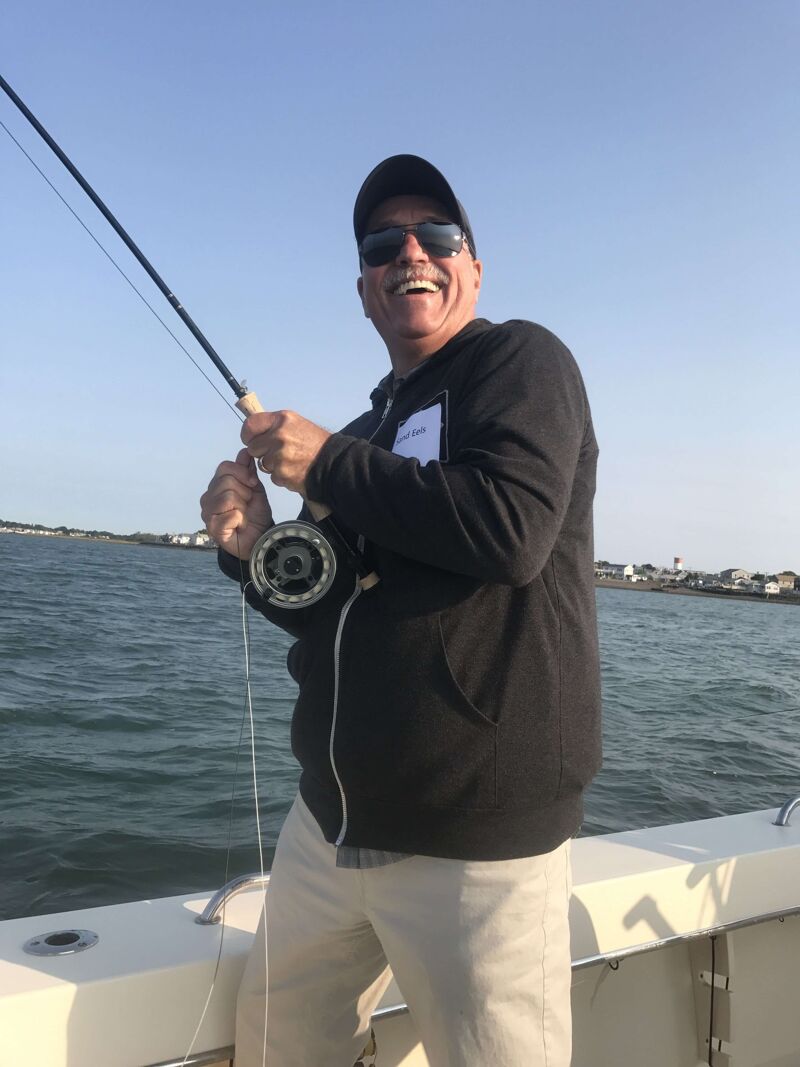 A man is smiling while fishing on a boat. He is wearing a black hoodie, khaki pants, a baseball cap, and sunglasses. The man is holding a fishing rod with a reel, and the water is calm with a clear blue sky above. The boat appears to be near the shore, with houses visible in the background.