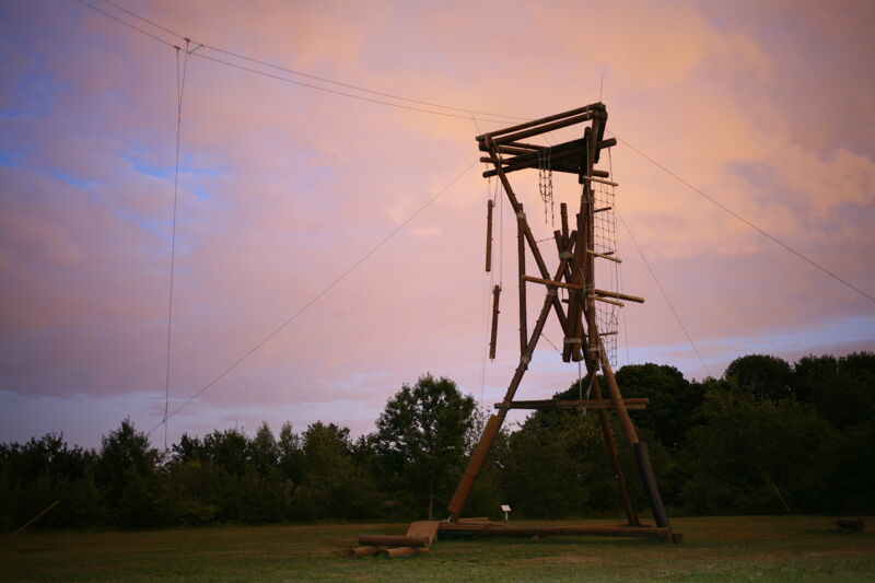 The image shows a wooden structure, possibly part of an adventure course, set against a colorful sunset sky. The structure has ropes, ladders, and hanging elements. It is located in a grassy area with trees in the background. The overall scene suggests an outdoor recreational setting.