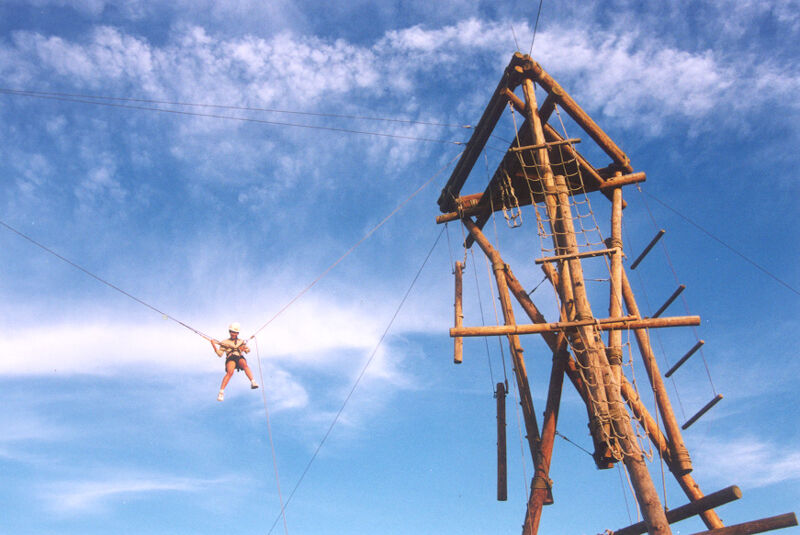 The image shows a person suspended in the air, likely engaged in a bungee jumping or similar activity. They are connected to a cable that extends towards a tall wooden structure. The sky is blue with some clouds. The wooden structure appears to be a tower with various platforms and supports.