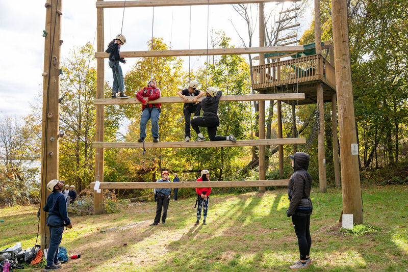 The image shows a group of people at an outdoor adventure course. Several individuals are climbing a wooden structure with ropes, while others are observing. The setting appears to be a park or wooded area, with trees and a lake visible in the background. The weather seems pleasant, with a clear sky.