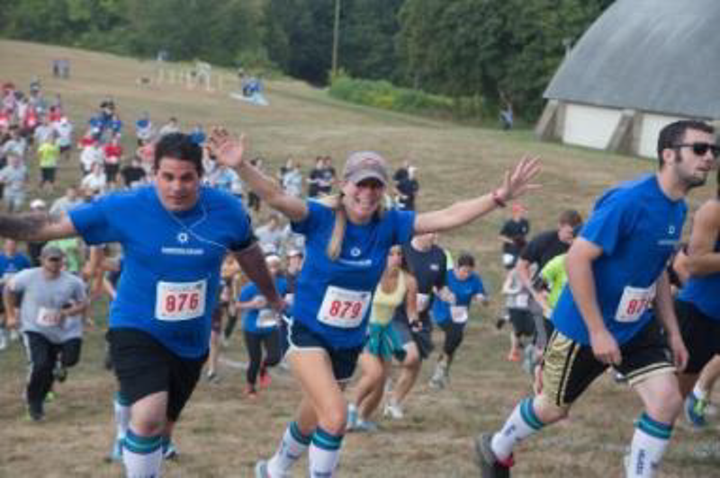 The image shows a group of people participating in a race. They are running uphill, wearing blue shirts with race numbers. The runners appear to be enjoying themselves, with some raising their arms in the air. The background includes a grassy hill and a building.