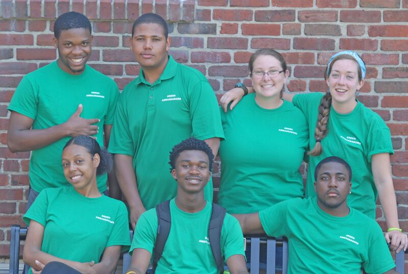 The image shows a group of seven people, all wearing green shirts, posing in front of a brick wall. They appear to be a team or a group of volunteers. Some are smiling, and the overall atmosphere seems positive and friendly.
