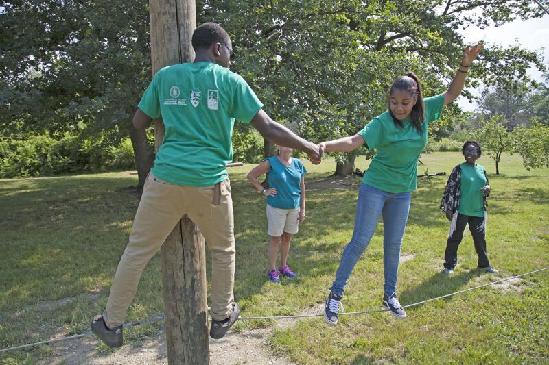 The image shows a group of people participating in an outdoor activity. Two individuals are actively engaged in a balancing act, with one person on a pole and another walking on a tightrope. Other people are watching them. They are all wearing green shirts. The setting appears to be a park or recreational area.