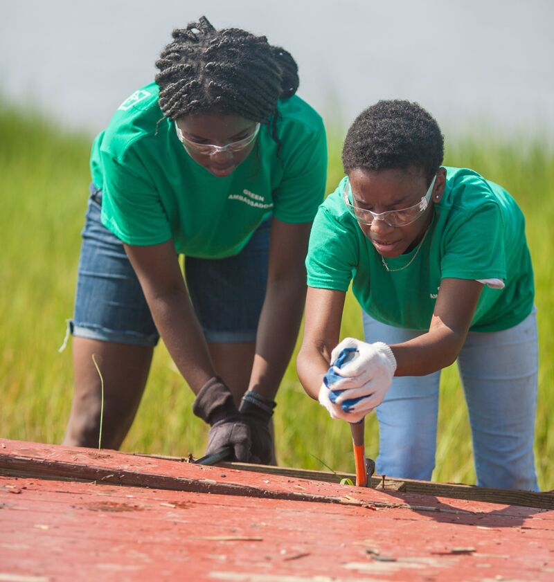 The image shows two young women working together outdoors. They are wearing green shirts, safety glasses, and gloves, suggesting they are engaged in a hands-on project. They appear to be focused on a wooden structure, possibly repairing or constructing something. The background features green grass, indicating an outdoor setting.