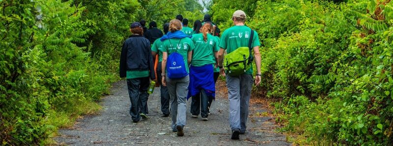 The image shows a group of people walking on a path surrounded by lush green trees. They appear to be on a hike or a nature walk. Most of them are wearing green shirts, suggesting they might be part of a group or organization. The path is made of gravel, and the overall scene conveys a sense of outdoor activity and nature.