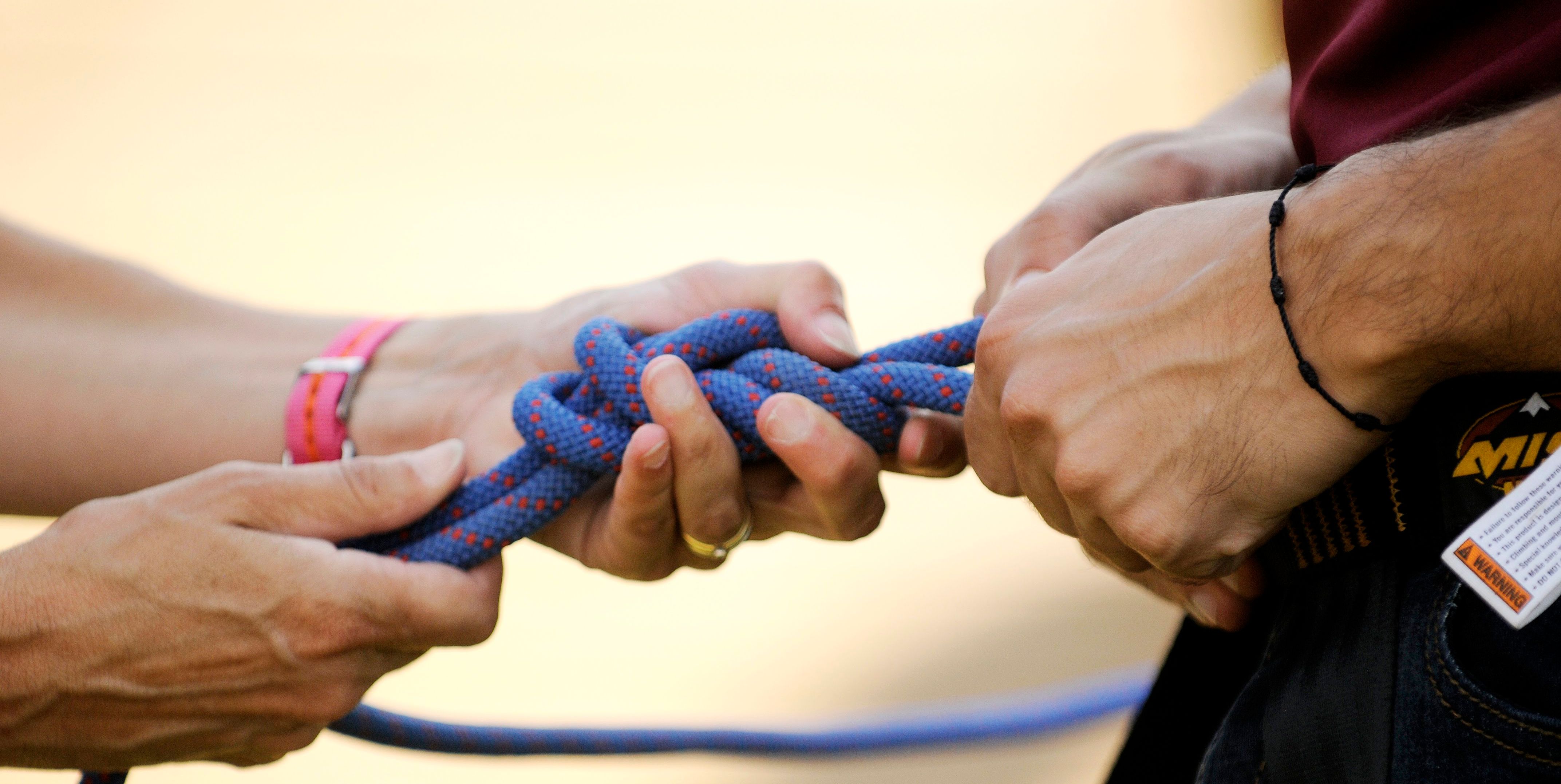 The image shows two people's hands tying a blue rope into a knot. The hands are in the process of securing the knot, with the rope forming loops and twists. The focus is on the intricate details of the knot-tying process, highlighting the precision and skill involved in creating a secure connection.