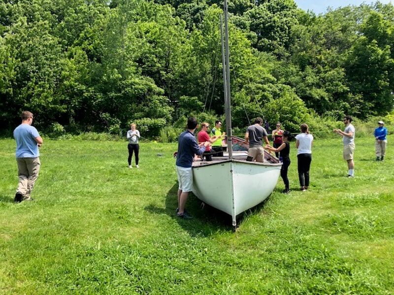 A group of people are gathered around a small sailboat on a grassy field, with a dense forest in the background. Some individuals appear to be preparing the boat, possibly for a sailing lesson or an event. The weather seems pleasant, with ample sunlight filtering through the trees, creating a serene outdoor atmosphere.