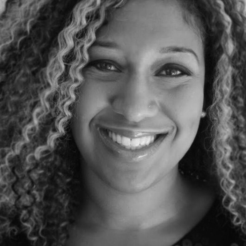 This is a black and white close-up photograph of a smiling woman with curly hair. She has a warm and friendly expression, with her eyes crinkled at the corners. The lighting highlights her features, creating a sense of depth and dimension. The overall impression is one of joy and approachability.