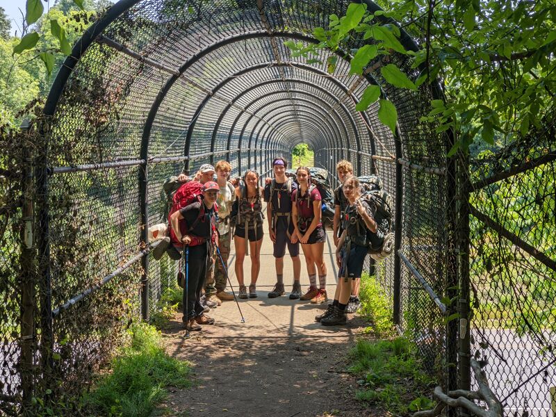 A group of hikers stands inside a long, arched tunnel made of metal mesh. They are wearing backpacks and hiking gear, suggesting they are on an outdoor adventure. The tunnel is surrounded by lush greenery, with trees and plants visible on either side. The lighting is bright, indicating it is daytime.