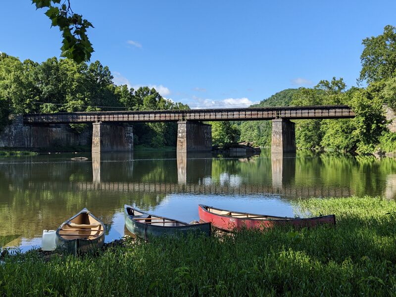 The image shows a serene river scene with three canoes resting on the grassy bank in the foreground. A long, dark bridge stretches across the river in the background, supported by stone pillars. The water reflects the bridge and sky, creating a symmetrical effect. Lush green trees line both sides of the river, adding to the peaceful and natural setting under a clear blue sky.